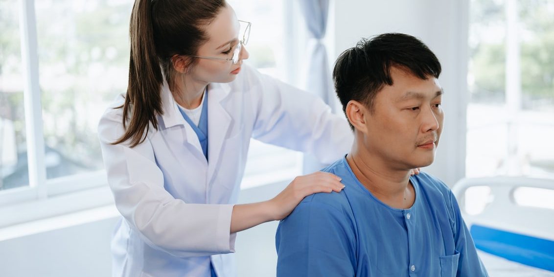 Female physiotherapist in lab coat examining the back of a male patient seated in a clinic.