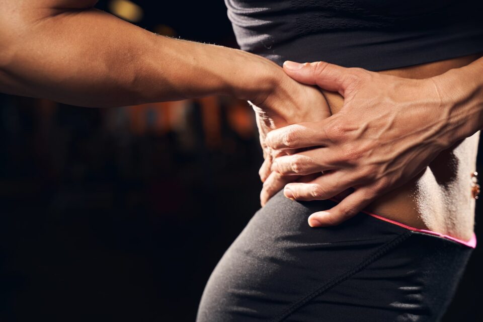 Close-up of a woman’s hands on her lower back, indicating pain, with a blurred gym background. Close-up of a woman's hands on her lower back, indicating pain, with a blurred gym background.