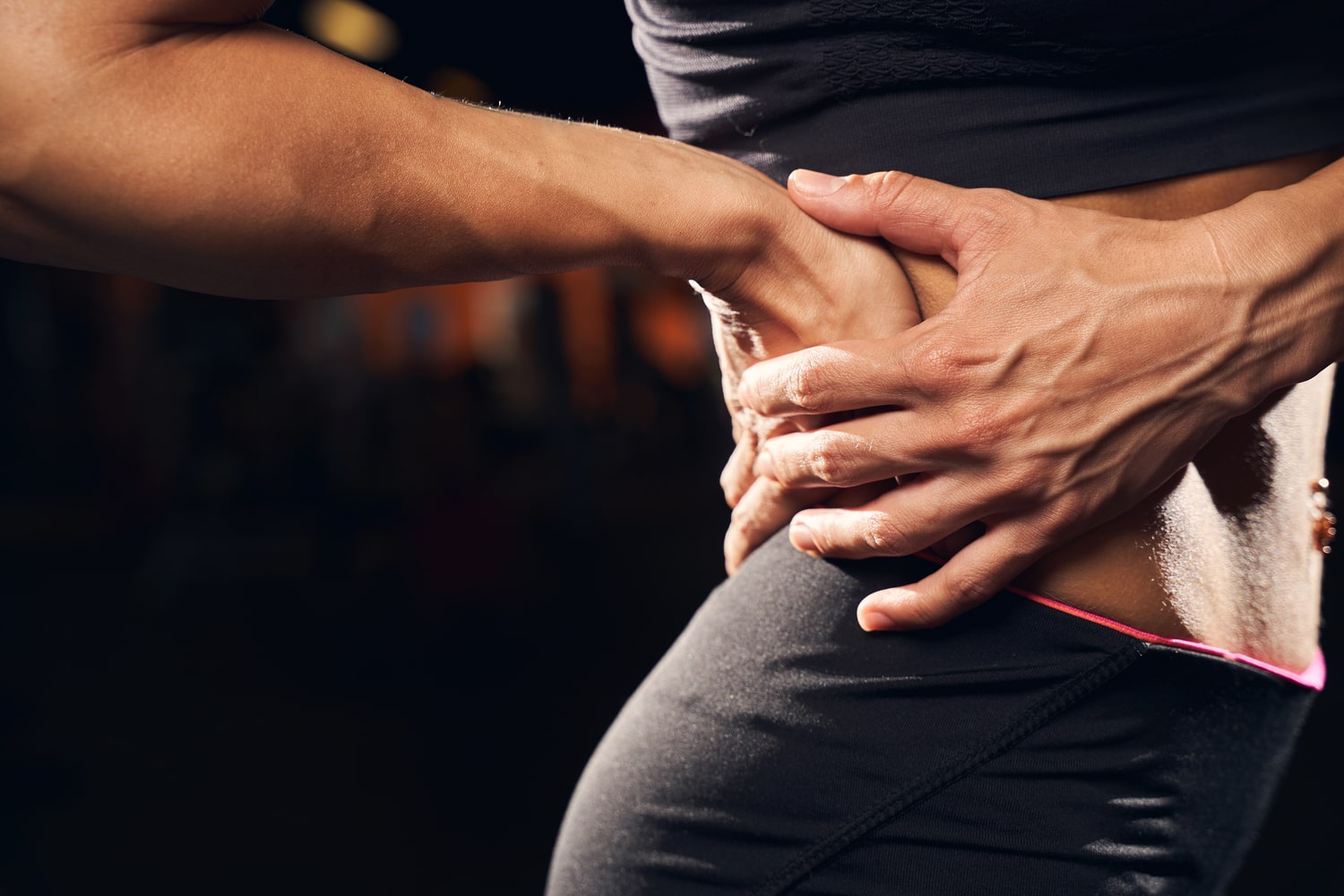 Close-up of a woman’s hands on her lower back, indicating pain, with a blurred gym background. Close-up of a woman's hands on her lower back, indicating pain, with a blurred gym background.