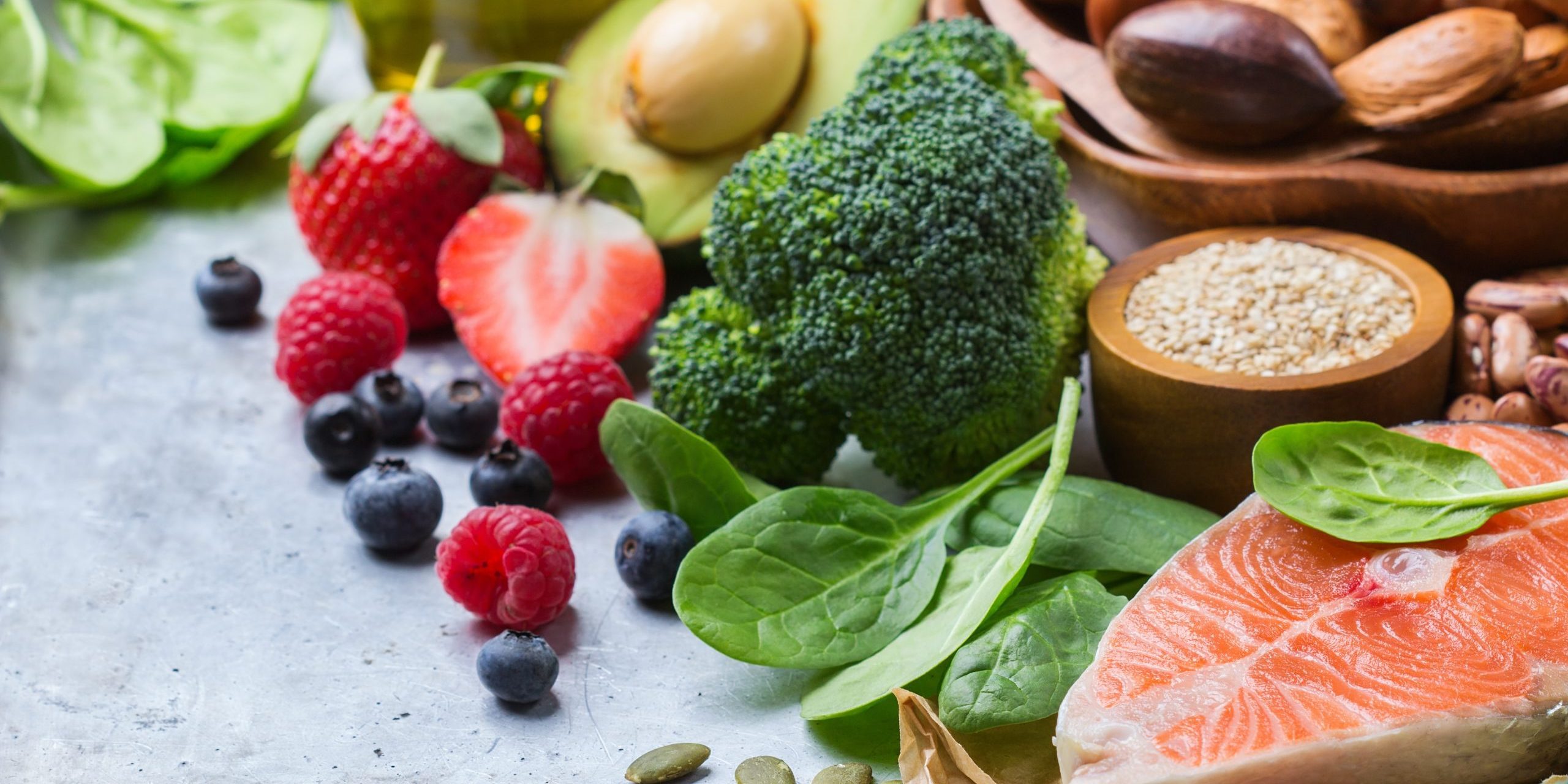 Assortment of healthy foods including salmon, nuts, seeds, berries, and vegetables on a light surface.