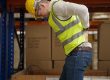 A warehouse worker in a yellow vest and helmet suffering from lower back pain while working.
