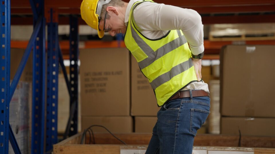 A warehouse worker in a yellow vest and helmet suffering from lower back pain while working. A warehouse worker in a yellow vest and helmet suffering from lower back pain while working.
