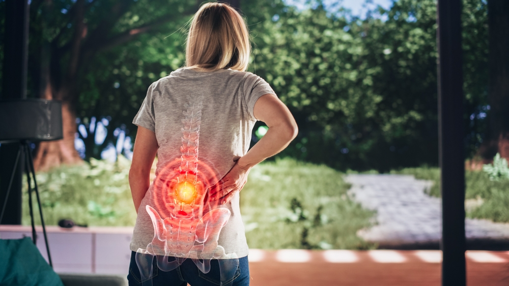 A person stands outdoors, holding their lower back in pain. An anatomical illustration of the spine highlights a red area in the lower back, indicating discomfort or injury. A person stands outdoors, holding their lower back in pain. An anatomical illustration of the spine highlights a red area in the lower back, indicating discomfort or injury.