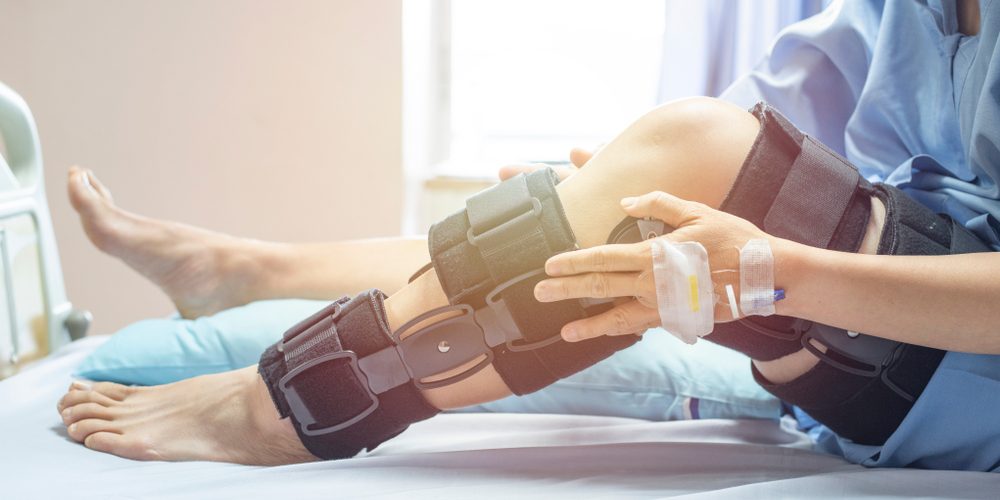 A person with a hospital gown and IV in their hand rests in bed while wearing a large knee brace on one leg. Pain Management for Post-Surgical Recovery
