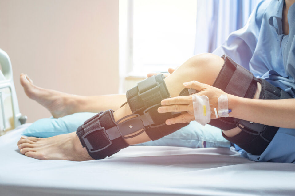 A person with a hospital gown and IV in their hand rests in bed while wearing a large knee brace on one leg. Pain Management for Post-Surgical Recovery