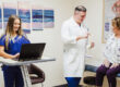 A healthcare provider in a white coat shows a spine model to a patient sitting on an exam table, while discussing Interventional Pain Management Techniques. Meanwhile, a nurse in scrubs types on a laptop.