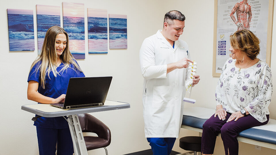A healthcare provider in a white coat shows a spine model to a patient sitting on an exam table, while discussing Interventional Pain Management Techniques. Meanwhile, a nurse in scrubs types on a laptop.