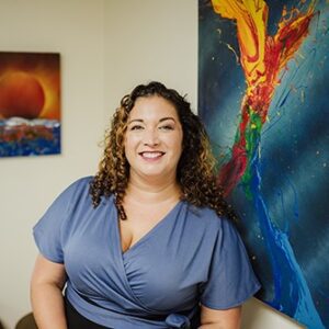 Woman with curly hair wearing a blue blouse stands smiling in front of colorful abstract paintings on a wall.