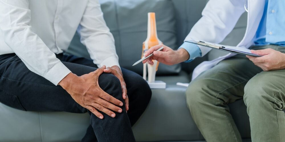 A person holds their knee while sitting on a couch. A doctor next to them points to a knee model and holds a clipboard. Holistic Pain Management Venice Florida