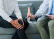 A person holds their knee while sitting on a couch. A doctor next to them points to a knee model and holds a clipboard. Holistic Pain Management Venice Florida