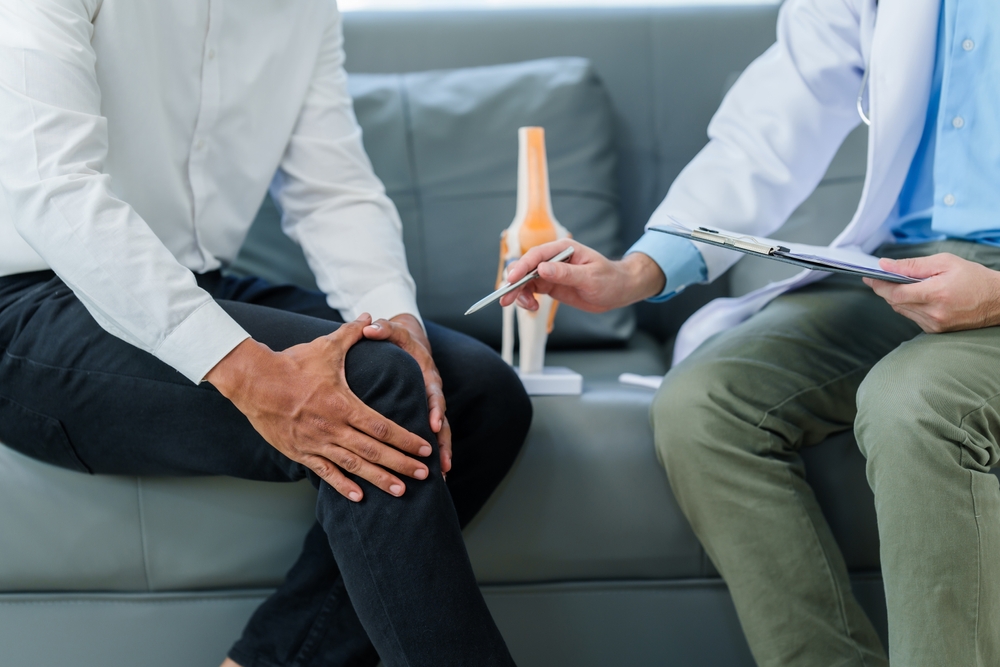 A person holds their knee while sitting on a couch. A doctor next to them points to a knee model and holds a clipboard. A person holds their knee while sitting on a couch. A doctor next to them points to a knee model and holds a clipboard. Holistic Pain Management Venice Florida