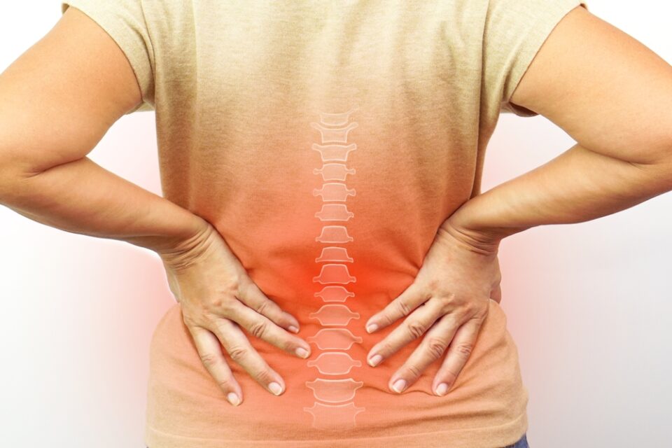 A person holds their lower back with both hands; an illustration of a spine is superimposed, highlighting the lower back area in red.