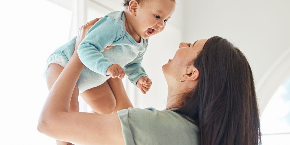 A woman smiles and lifts a happy baby wearing a light blue onesie in a bright room.