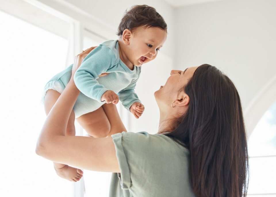 A woman smiles and lifts a happy baby wearing a light blue onesie in a bright room. A woman smiles and lifts a happy baby wearing a light blue onesie in a bright room.