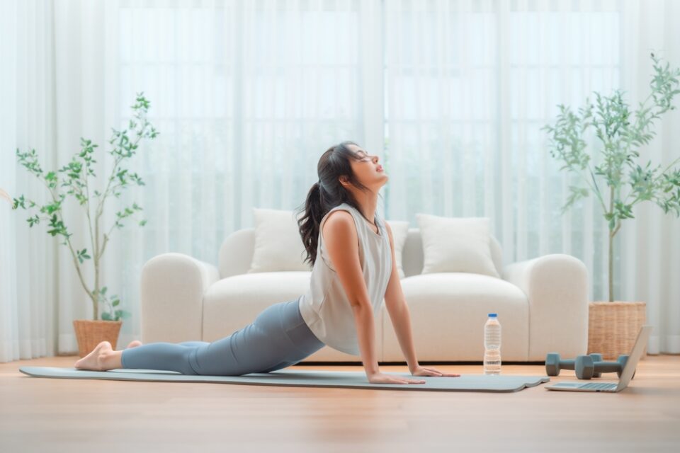 A woman practices yoga indoors, performing an upward-facing dog pose on a mat with a water bottle and dumbbells nearby. spine health exercises venice fl, port charlotte fl