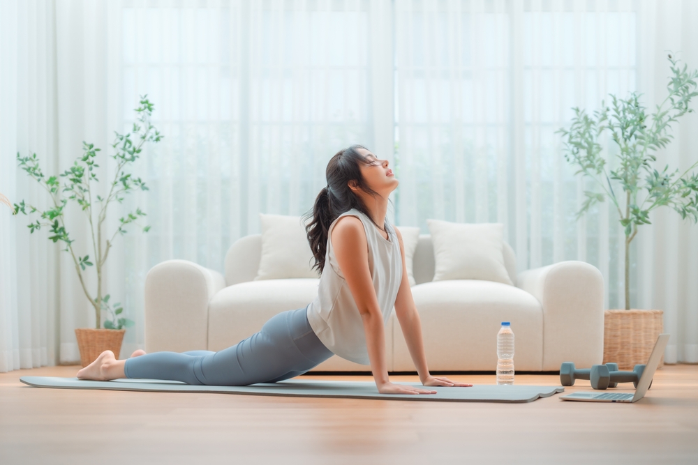 A woman practices yoga indoors, performing an upward-facing dog pose on a mat with a water bottle and dumbbells nearby. spine health exercises venice fl, port charlotte fl