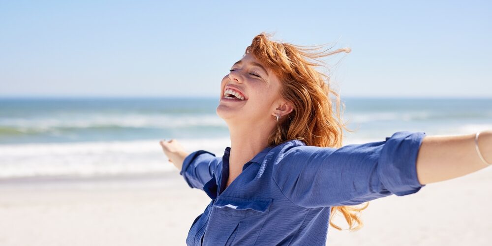 A woman with red hair smiles with outstretched arms on a sandy beach with the ocean and blue sky in the background. Chronic Joint Pain
