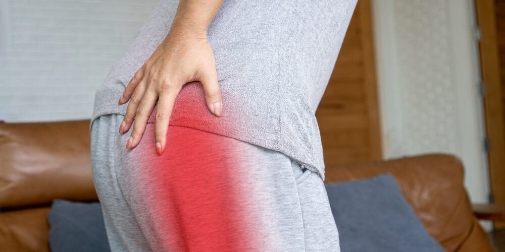A person holding their lower back and hip area, with a red highlight indicating pain or discomfort, standing in a living room. Sciatica Pain