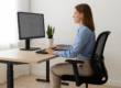 A woman sitting upright with perfect posture at a modern ergonomic desk in a bright, minimalist office, using a computer monitor positioned at eye level — illustrating healthy workspace posture for back and neck pain prevention.