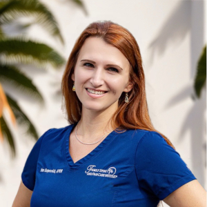 A woman with long red hair wearing blue medical scrubs stands outdoors, smiling at the camera; palm fronds and a white wall are in the background.