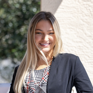 A woman with long blonde hair, wearing a patterned top and black blazer, smiles while standing outdoors near a light-colored wall.