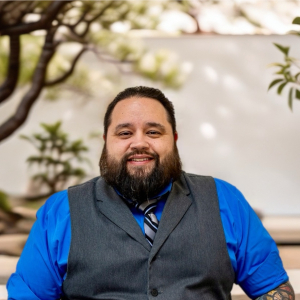A man with a beard, wearing a blue shirt, gray vest, and striped tie, sits outdoors in front of trees and a white wall, smiling at the camera.