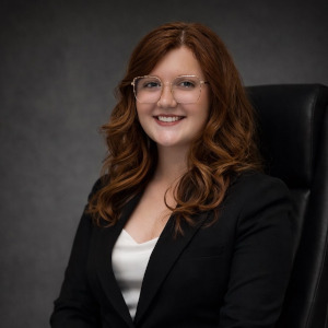 A woman with long red hair and glasses, wearing a black blazer and white top, sits on a black chair against a dark background.