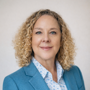 A woman with curly blonde hair wearing a light blue blazer and a white patterned shirt, smiling at the camera against a plain background.