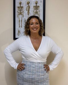 Adeline stands smiling in front of a framed anatomical skeleton chart on a beige wall, wearing a white long-sleeve top and a gray plaid skirt.