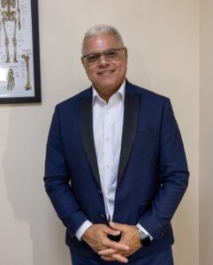 A man with white hair and glasses, wearing a blue suit jacket and white shirt, stands smiling in an office with a skeletal anatomy chart on the wall.