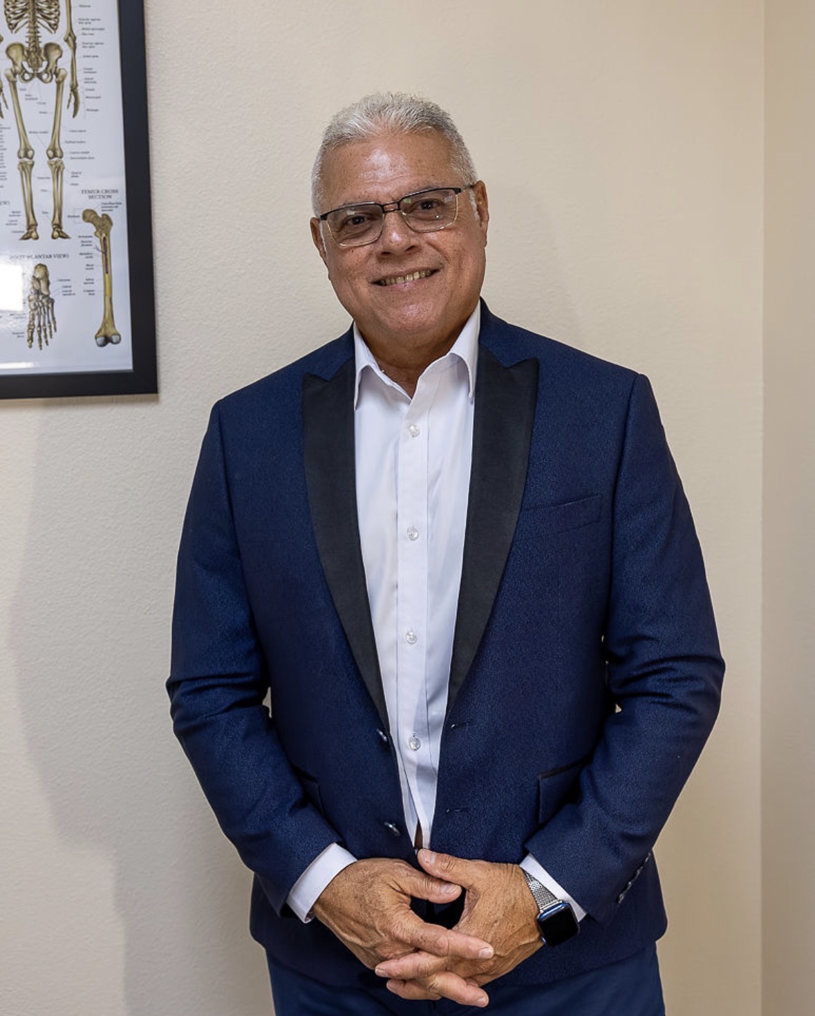 A man with white hair and glasses, wearing a blue suit jacket and white shirt, stands smiling in an office with a skeletal anatomy chart on the wall.