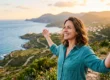 Woman smiling with arms outstretched on a coastal hillside at sunset, with a view of the ocean, winding roads, and distant mountains. smiling and experiencing migraine relief.