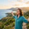 Woman smiling with arms outstretched on a coastal hillside at sunset, with a view of the ocean, winding roads, and distant mountains. smiling and experiencing migraine relief.