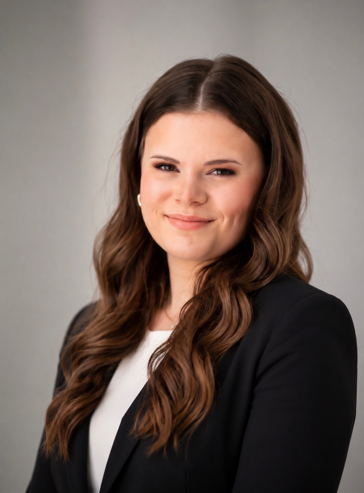 A staff member with long brown hair wearing a black blazer and white shirt smiles slightly in a professional headshot against a plain background.