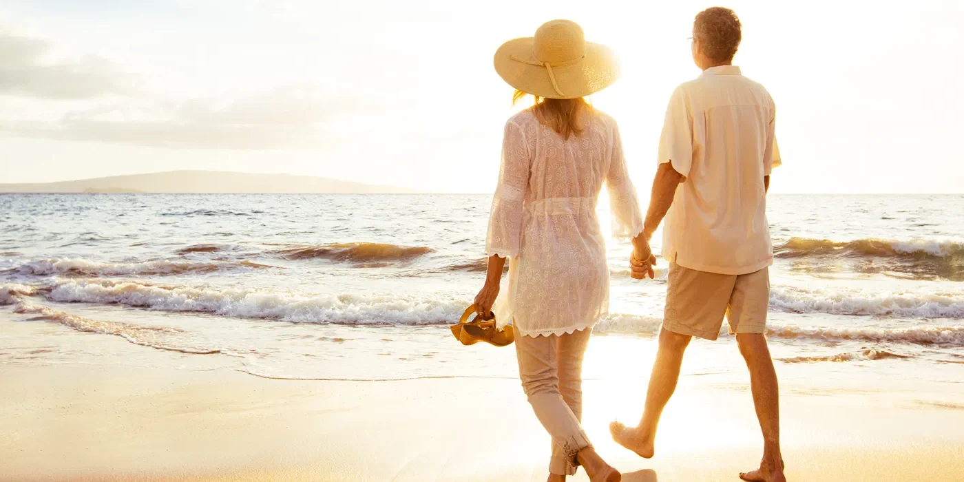 A couple walks hand in hand along a sandy beach at sunset, with gentle waves in the background. An older couple walks hand-in-hand along the shoreline at sunset, symbolizing spinal stenosis pain relief and renewed quality of life.