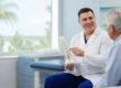 A doctor in a white coat holds a spine model and speaks to an older male patient in a medical office about Kyphoplasty.