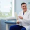 A doctor in a white coat holds a spine model and speaks to an older male patient in a medical office about Kyphoplasty.