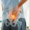 A person holds their lower back in SI joint pain; a digital illustration of the spine and pelvis highlights a painful area.